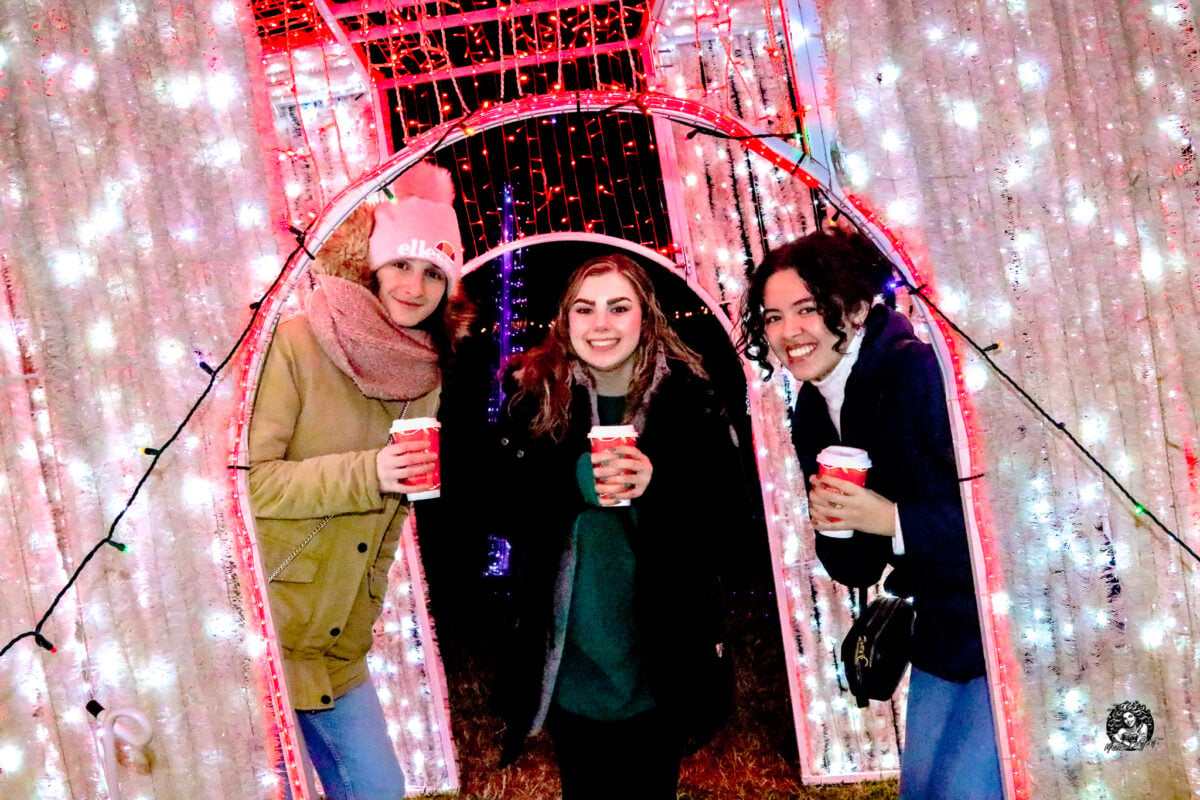Three friends with hot drinks in a tunnel of holiday lights